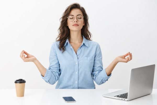 Girl meditating at work sitting in lotus pose with nirvana orbs near laptop and smartphone trying relax and get rid of pressure and stress posing calm and peaceful against gray background in glasses